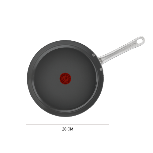 Na obrazie widoczna jest patelnia z ciemnym wnętrzem i metalowym uchwytem. Na środku patelni znajduje się czerwony okrągły symbol.