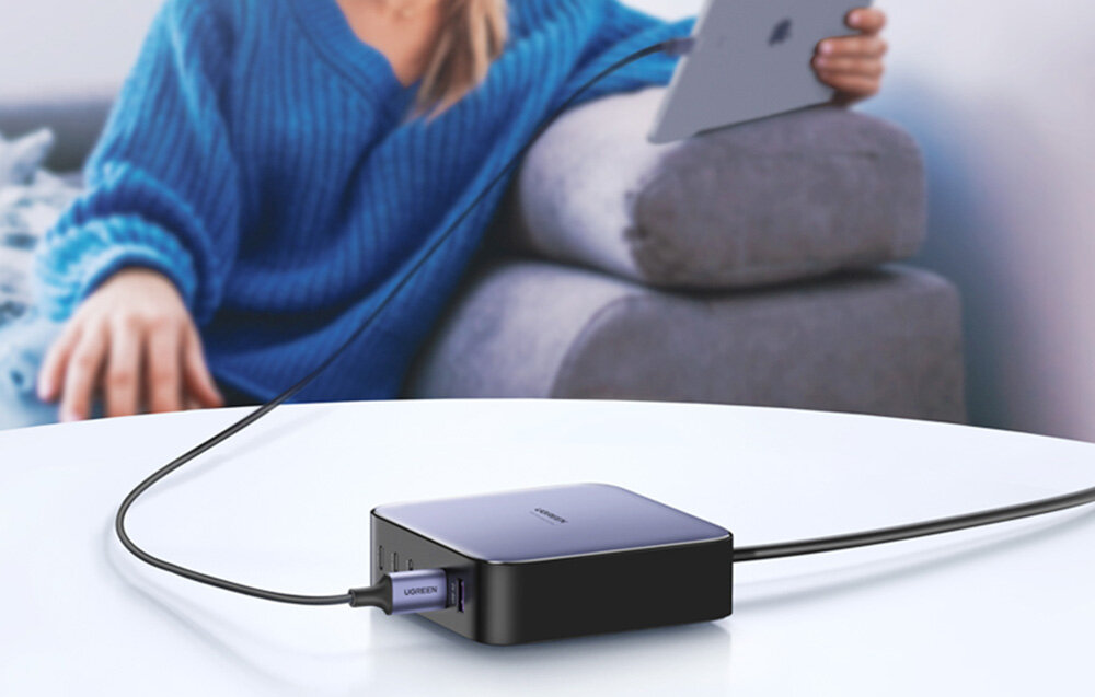Na białym stole leży czarna ładowarka z kilkoma portami USB, do której podłączony jest kabel. W tle osoba siedzi na kanapie, trzymając tablet.