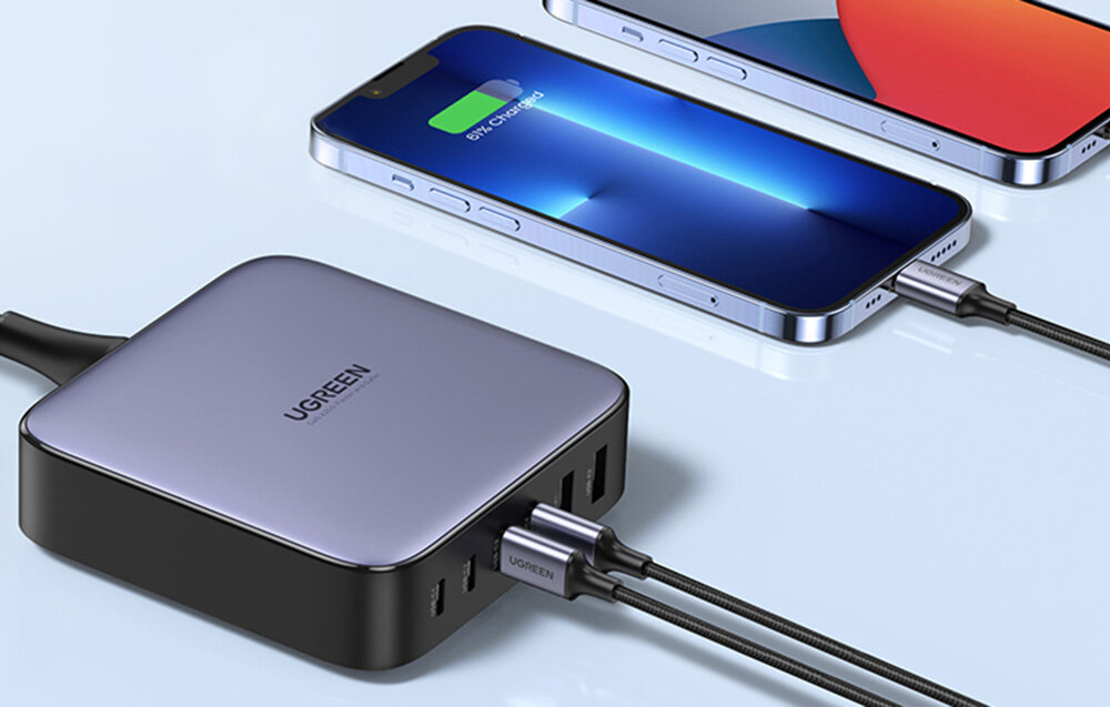 Na obrazie widoczna jest czarna ładowarka z logo UGREEN, podłączona do dwóch smartfonów. Ładowarka posiada kilka portów USB, a jeden z telefonów pokazuje ikonę ładowania baterii.