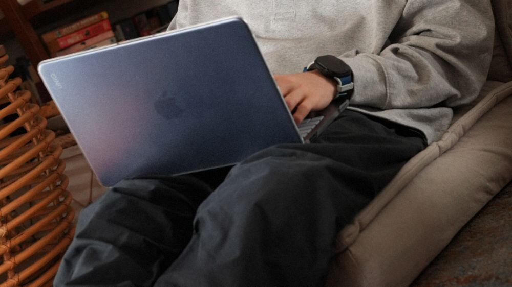 Osoba siedzi na kanapie, trzymając laptopa w przezroczystym etui. Na etui widoczne jest logo producenta w górnym rogu.