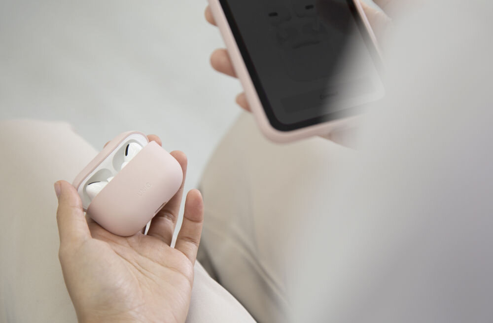 Osoba trzyma różowe etui na słuchawki, w którym znajdują się słuchawki. W tle widoczny jest telefon w różowym etui.