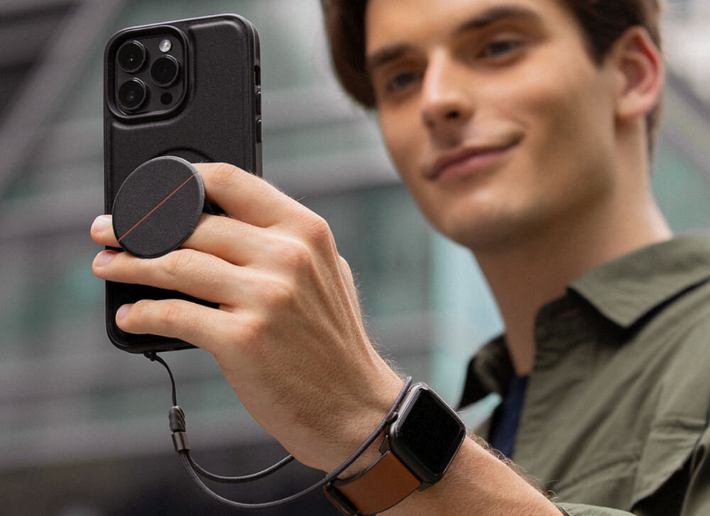 Osoba trzyma smartfon z czarnym etui, do którego przymocowany jest okrągły uchwyt. Na nadgarstku osoby znajduje się smartwatch z brązowym paskiem.