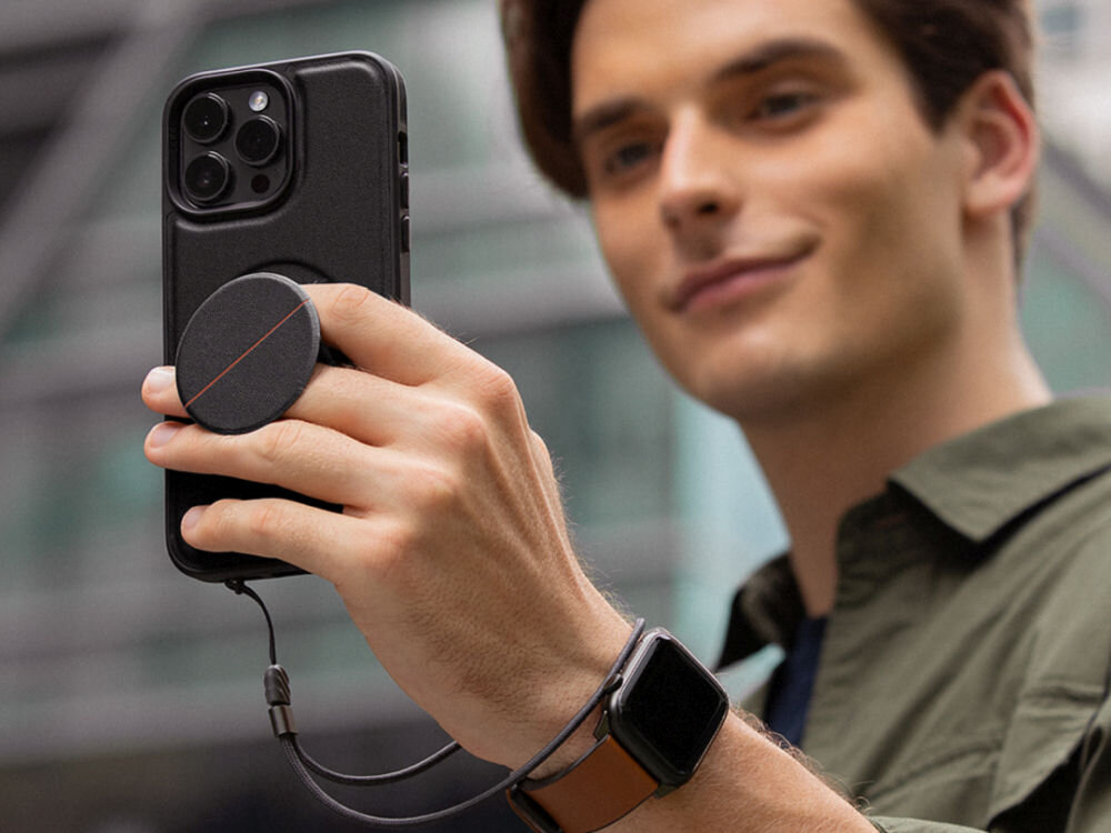 Osoba trzyma smartfon z czarnym etui, na którym zamocowany jest okrągły uchwyt. Na nadgarstku widoczny jest smartwatch z brązowym paskiem.