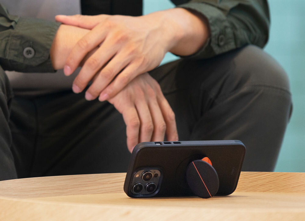 Na drewnianym stole leży telefon w czarnym etui z zamontowanym uchwytem. W tle widoczna jest osoba siedząca z rękami opartymi na kolanach.