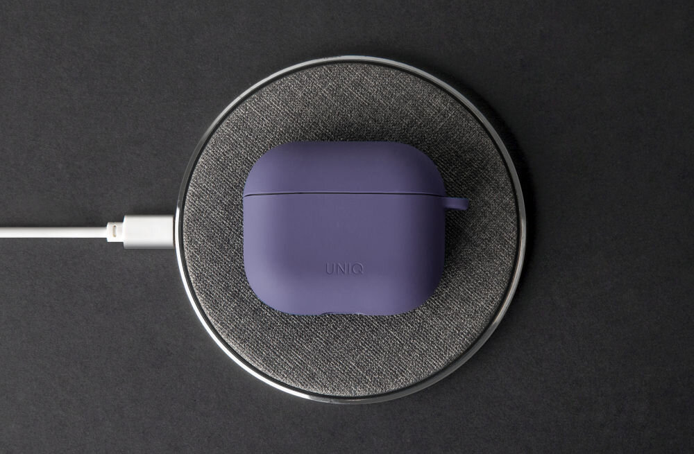 Purpurowe etui na słuchawki z napisem \'UNIQ\' leży na okrągłej, szarej podstawce do ładowania. Podstawka jest podłączona do kabla USB.