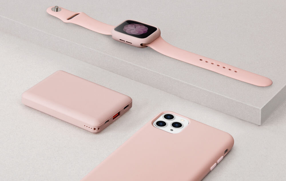 Na obrazie widoczne są różowe akcesoria elektroniczne: smartwatch, powerbank i smartfon. Wszystkie przedmioty mają jednolitą, pastelową kolorystykę.