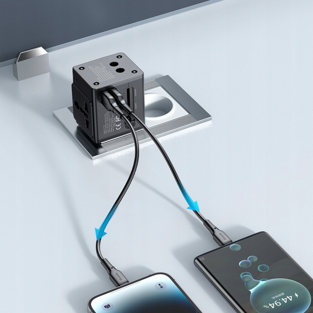 Na obrazie widać czarną ładowarkę USB podłączoną do gniazdka, z której wychodzą dwa kable ładujące dwa smartfony. Na ekranie jednego z telefonów widoczny jest wskaźnik ładowania baterii.
