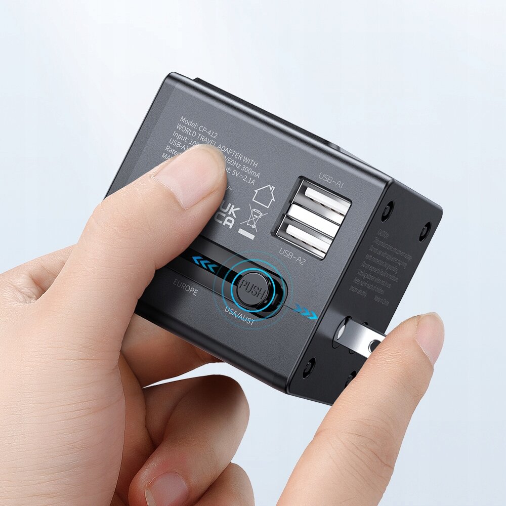 Osoba trzyma czarną ładowarkę USB z dwoma portami oznaczonymi jako USB-A1 i USB-A2 oraz przyciskiem z napisem \'PUSH\'. Widoczny jest tekst: \'Model: CR-412 WORLD TRAVEL ADAPTER WITH USB: 100-240V~50/60Hz 300mA USB-A: 5V-2.4A\'.