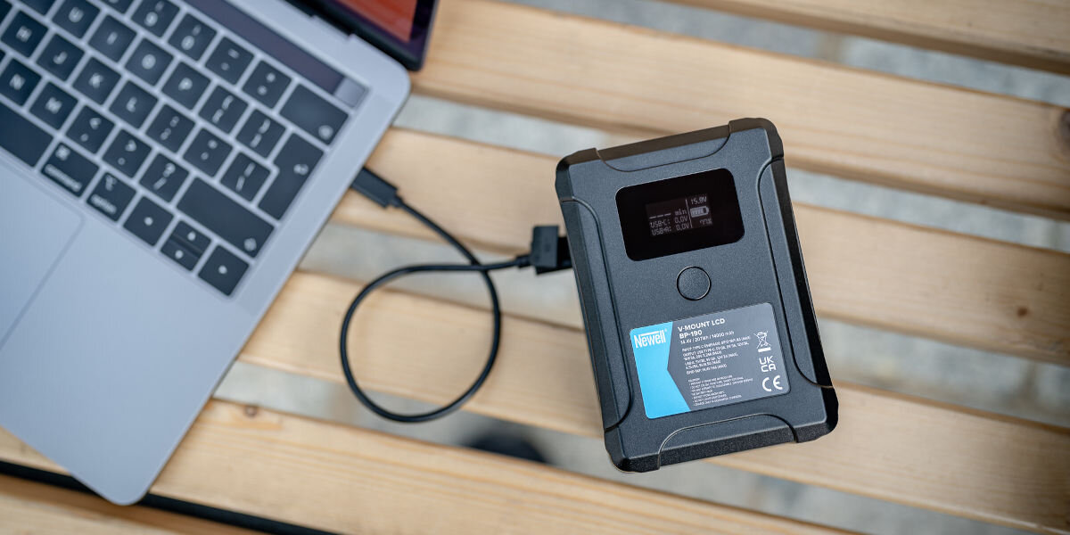 Akumulator NEWELL 14000 mAh do BP-190 LCD V-Mount, pojemnośc, ładowanie, wiele, porty, akumulator leży na deskach i ładuje obok laptopa
