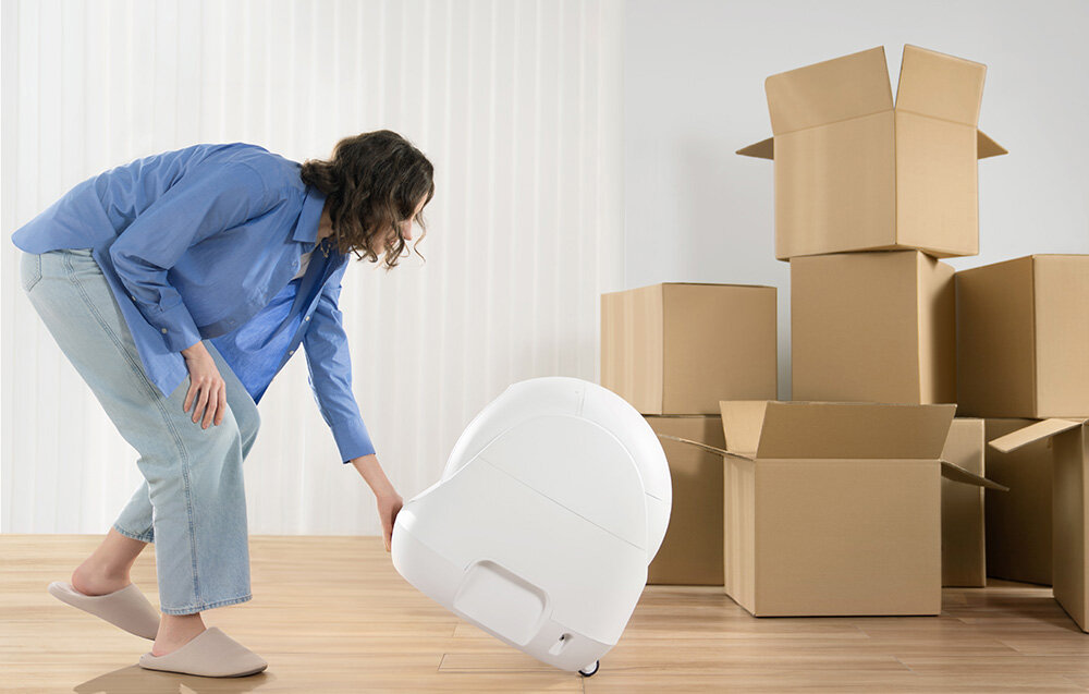Inteligentna kuweta dla kota CATLINK Baymax kobieta ustawia białą kuwetę obok leżą kartony kompaktowe wymiary (599 x 533 x 545 mm) elegancka minimalistyczna estetyka zintegrowane kółka przestronne wnętrze z nisko umieszczonym wejściem w każdym wieku o różnym stanie zdrowia komfort pupila