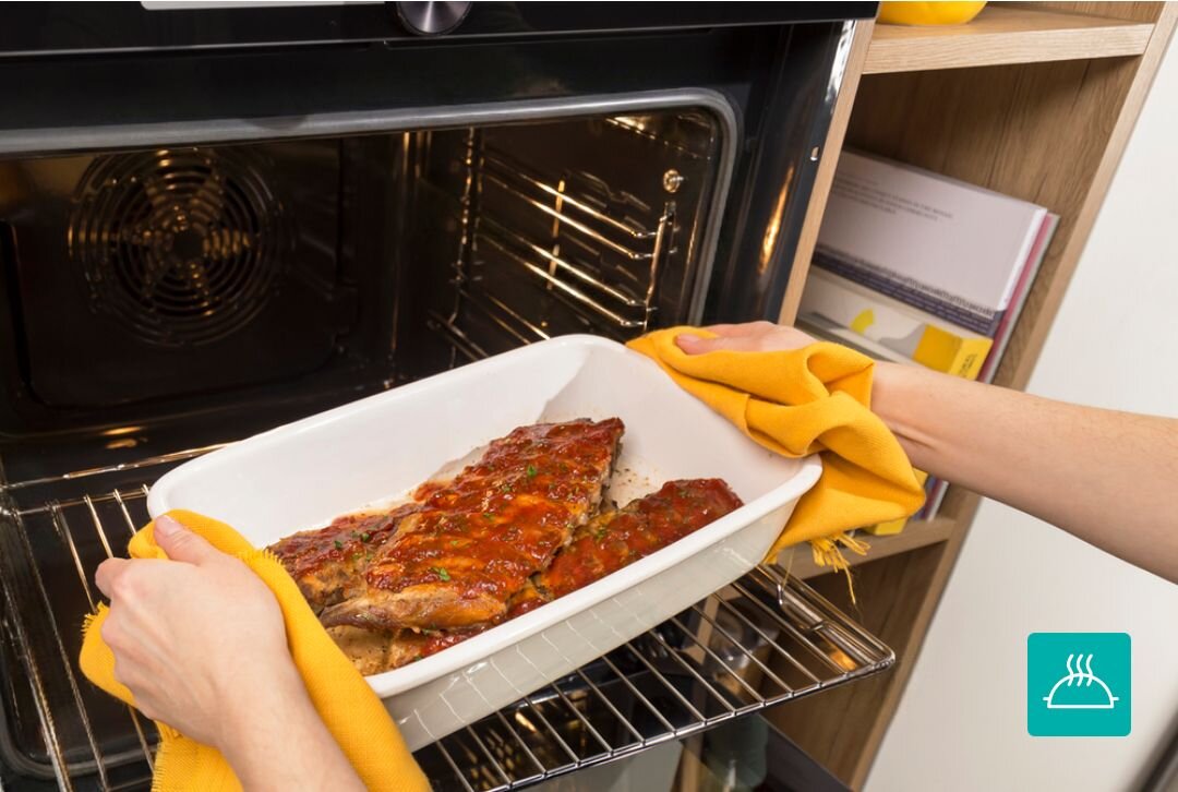 PIEKARNIK GORENJE BOS67371SYB Wyjmowanie gorącej potrawy w naczyniu żaroodpornym z piekarnika przy użyciu kuchennych rękawic. Zdjęcie akcentuje bezpieczeństwo i wygodę użytkowania