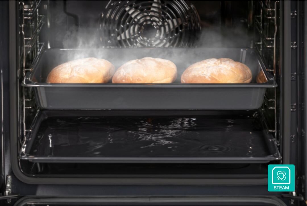 PIEKARNIK GORENJE BOS67371SYB Trzy bochenki chleba pieką się na blasze w piekarniku z aktywną funkcją pary, widoczną w postaci unoszącej się mgły. Ujęcie podkreśla równomierne wypiekanie i efekt chrupiącej skórki