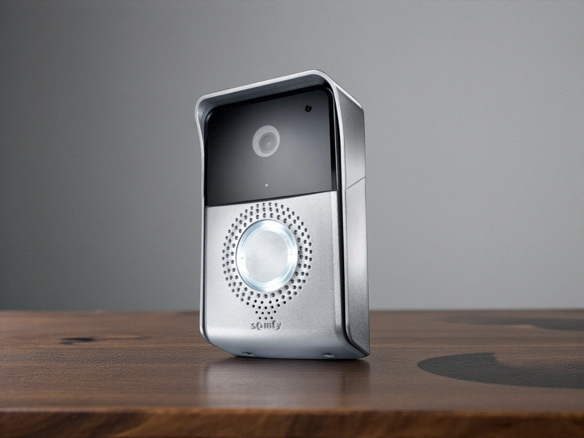 Wideodomofon SOMFY V500, personalizacja, historia, interkom, sterowanie, Panel zewnętrzny SOMFY V500 na drewnianym stole, zbliżenie na soczewkę kamery i głośnik.