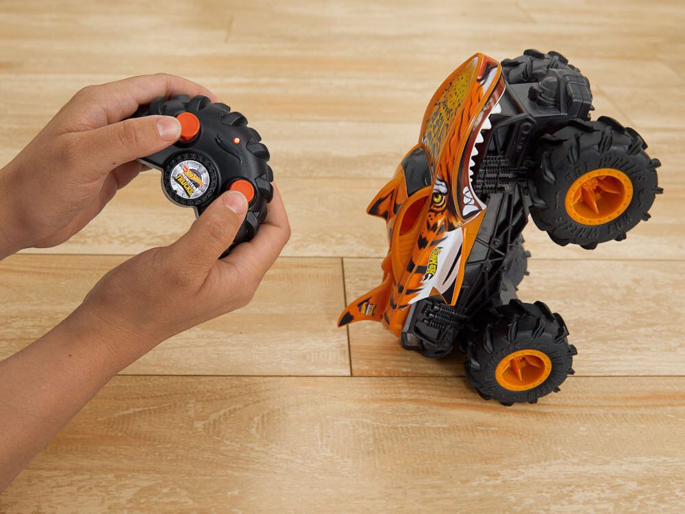 Samochód zdalnie sterowany Hot Wheels Tiger Shark HNV03 Dwie dłonie trzymające czarny kontroler (pilot). Obok, na drewnianej podłodze, zdalnie sterowany samochód Hot Wheels Monster Truck Shark przewrócony na bok, pokazujący podwozie i duże opony z pomarańczowymi felgami.