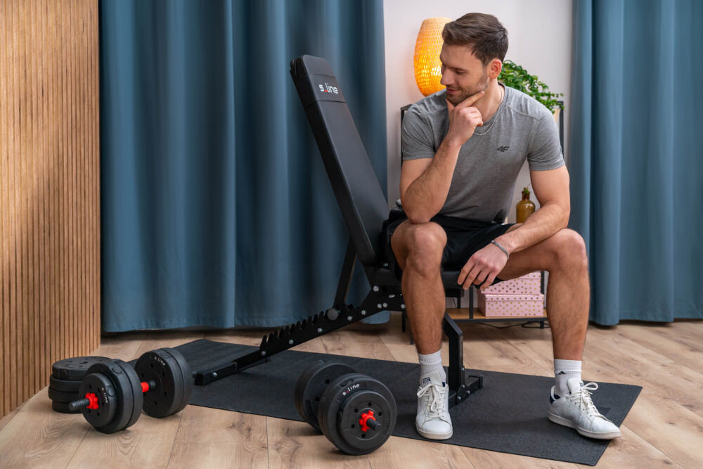 Ławka do ćwiczeń S-LINE ELLI BF300 Mężczyzna siedzący na regulowanej ławce do ćwiczeń S-line obok hantli na macie treningowej w domu.