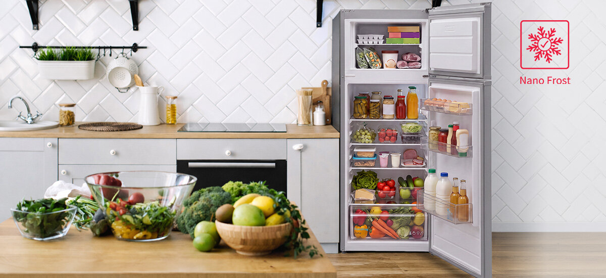 Otwarta lodówka z widocznym systemem Nano Frost, oznaczonym niebieskimi strzałkami wewnątrz. Obok lodówki znajduje się kuchnia z otwartą zmywarką i półką z roślinami oraz naczyniami.
