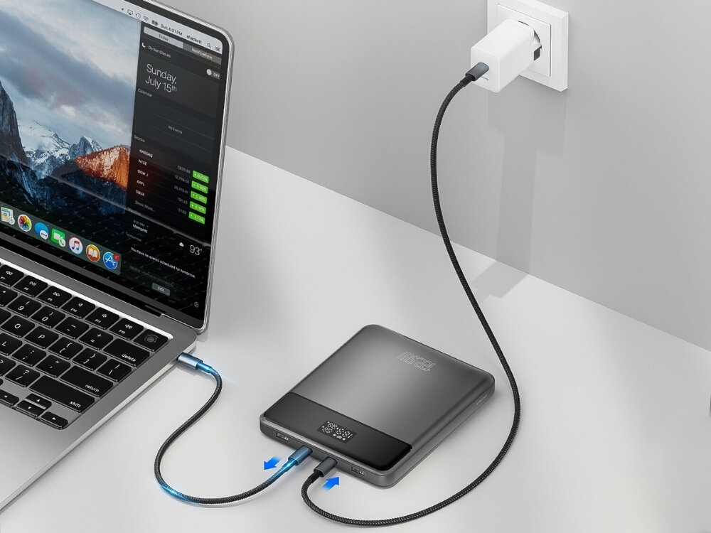 Na biurku znajduje się laptop podłączony do szarego powerbanku, który jest podłączony do gniazdka elektrycznego. Na ekranie laptopa widoczny jest krajobraz górski oraz data \'Sunday, July 15th\'.