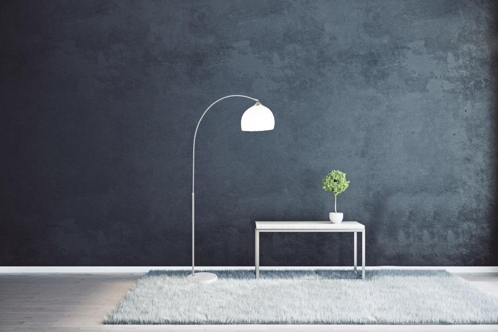 Na obrazie znajduje się lampa podłogowa z zakrzywionym ramieniem i białym kloszem, stojąca na dywanie. Obok lampy znajduje się stolik z małą rośliną w doniczce na tle ciemnej ściany.