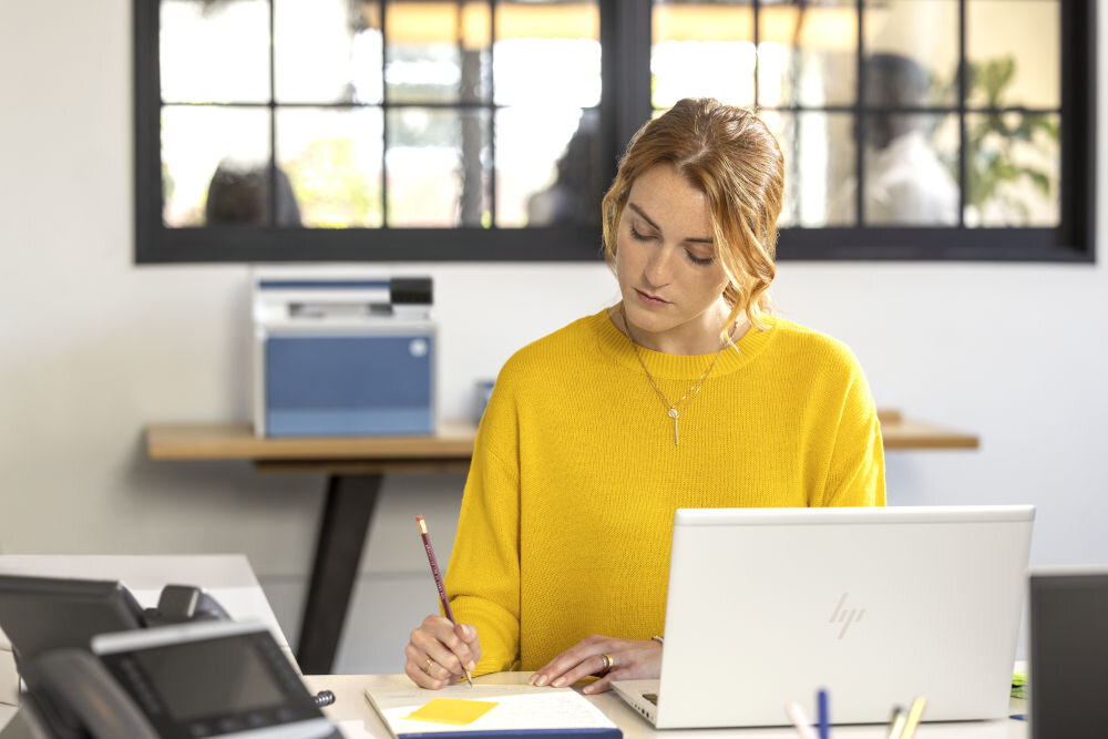 Urządzenie wielofunkcyjne HP Color LaserJet Pro 4302fdn Druk w kolorze, Automatyczny druk dwustronny prezentacja urządzenia w biurze w przodzie konieta pracująca przy laptopie siedząca przy biurku