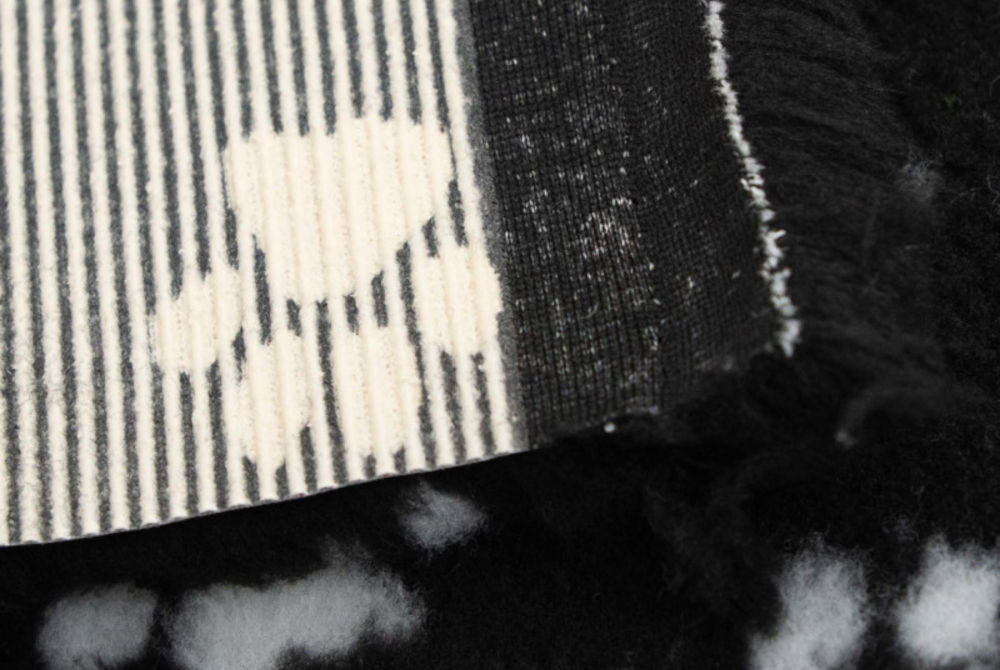 Legowisko uniwersalne BUBA Dry Bed A23 czarny w łapki 50 x 37 Zbliżenie na róg puszystego, czarnego dywanika. Wyraźnie widoczny jest spód z jasnej tkaniny w prążki oraz krawędź długiego, miękkiego włosia.