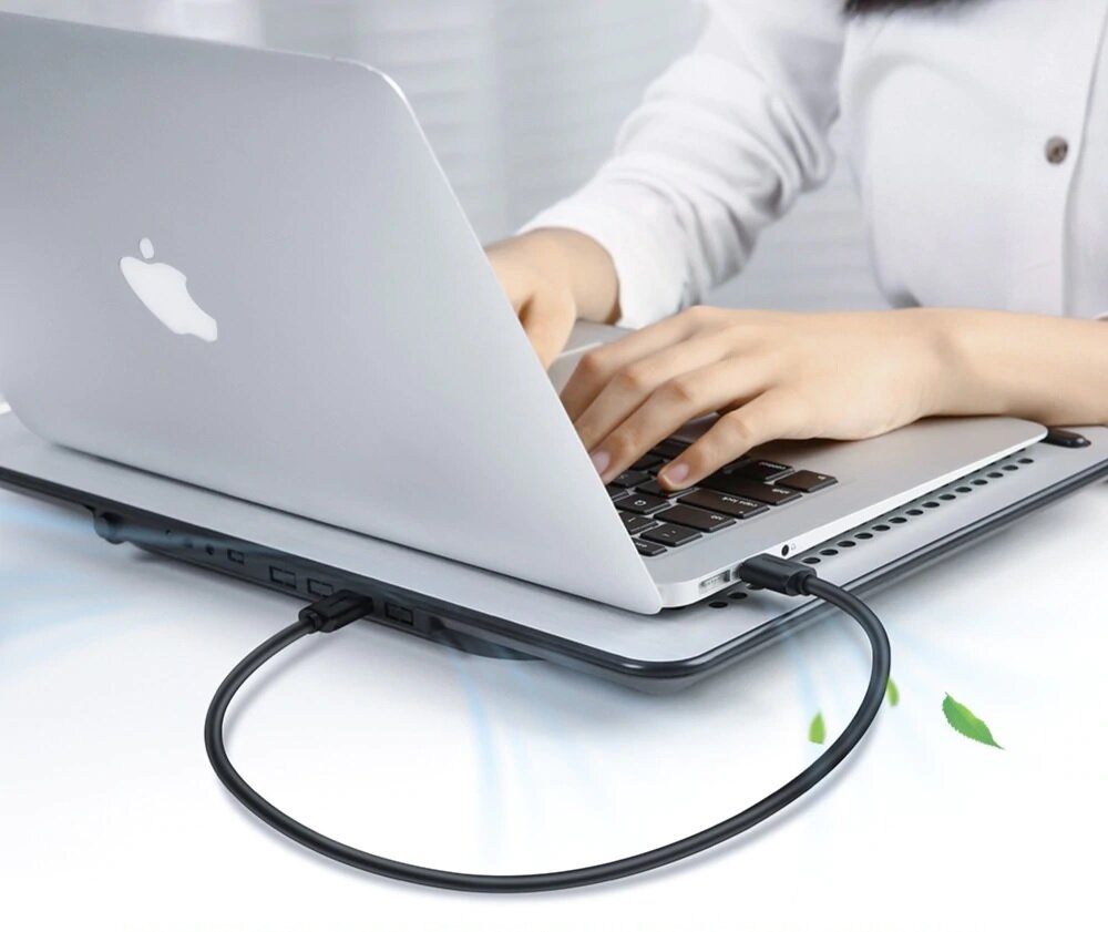 Osoba pisze na laptopie, do którego podłączony jest czarny kabel USB. Na obudowie laptopa widoczne jest logo w kształcie nadgryzionego jabłka.
