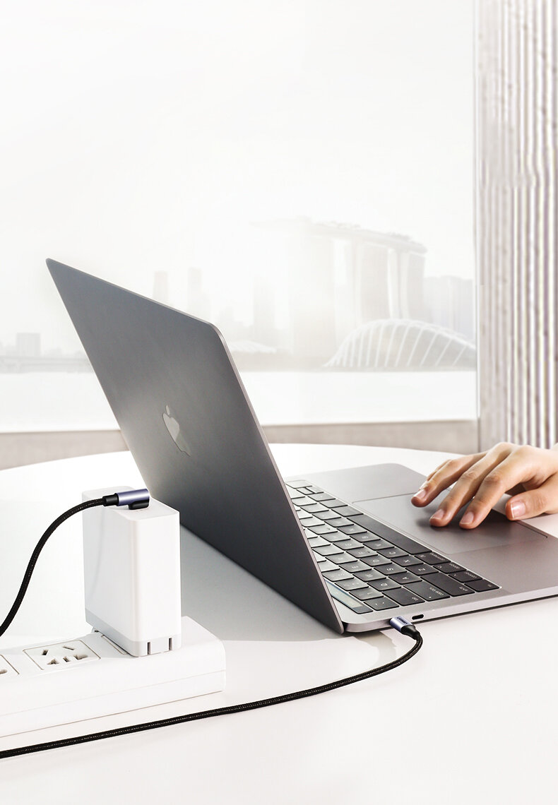 Na obrazie widać laptopa podłączonego do ładowarki za pomocą czarnego kabla USB-C. W tle znajduje się rozmyty widok miasta za oknem.