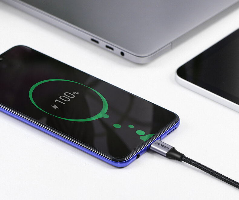 Na obrazie widać smartfon z wyświetlaczem pokazującym 100% naładowania baterii, podłączony do kabla USB-C z logo Ugreen. W tle znajduje się laptop i tablet.