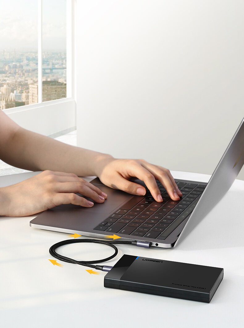 Osoba korzysta z laptopa, do którego podłączony jest czarny kabel USB-C z kątowym złączem. Kabel łączy laptop z czarnym urządzeniem z logo \'UGREEN\'.
