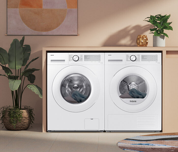 Na obrazie widoczne są dwie białe pralki ustawione obok siebie, z roślinami i dekoracjami w tle. Na jednej z pralek widnieje napis \'Heatpump\'.