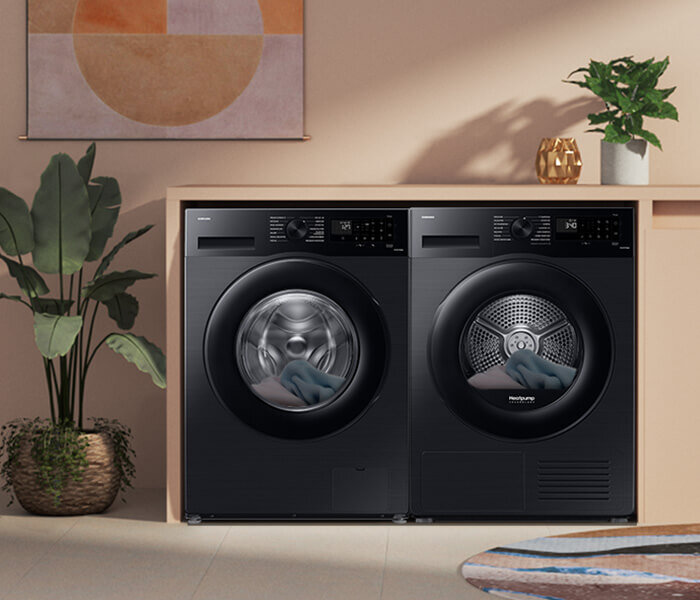 Na obrazie widoczne są dwie czarne pralki ustawione obok siebie, z otwartymi drzwiczkami, w których widać ubrania. W tle znajdują się rośliny doniczkowe i dekoracje ścienne.