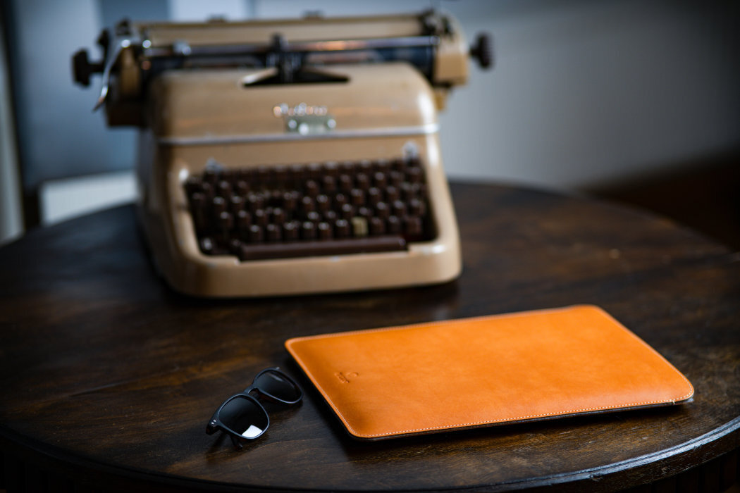 Na okrągłym drewnianym stole znajduje się brązowe etui na laptopa oraz okulary przeciwsłoneczne. W tle widoczna jest stara maszyna do pisania z logo \'Olivetti\'.