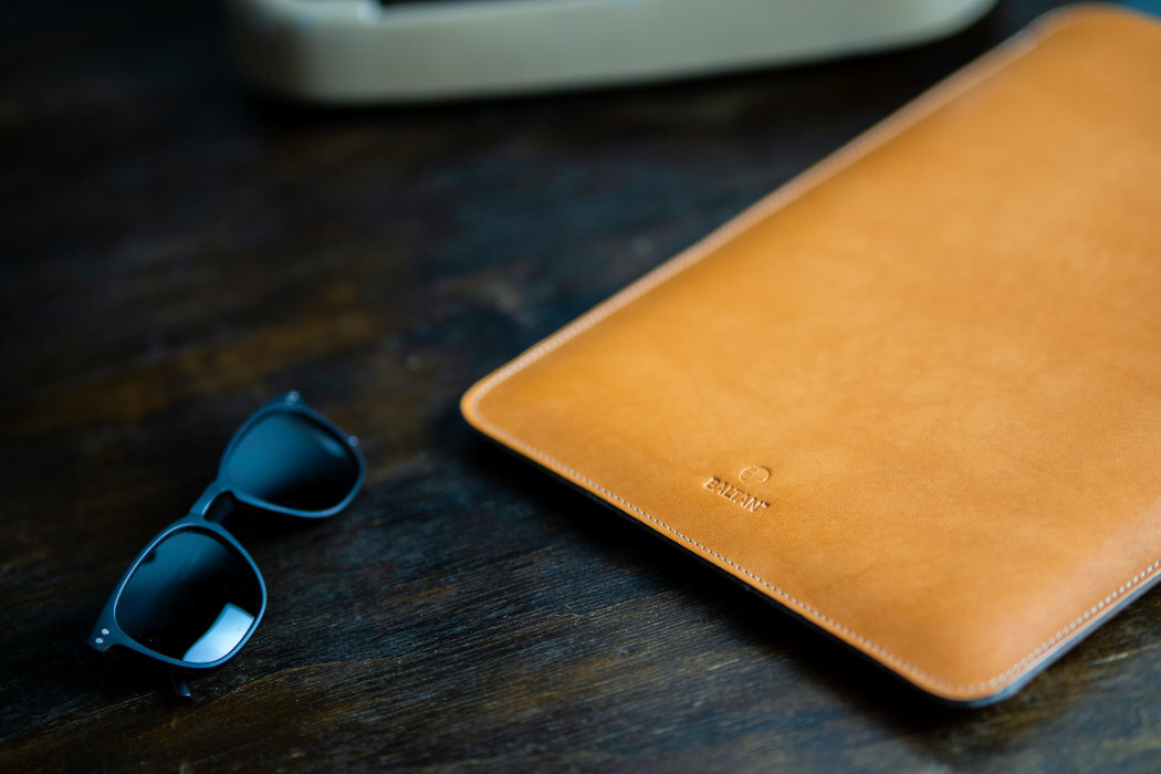 Brązowe etui na laptopa z logo \'BALTAN\' leży na ciemnym drewnianym stole obok czarnych okularów przeciwsłonecznych.
