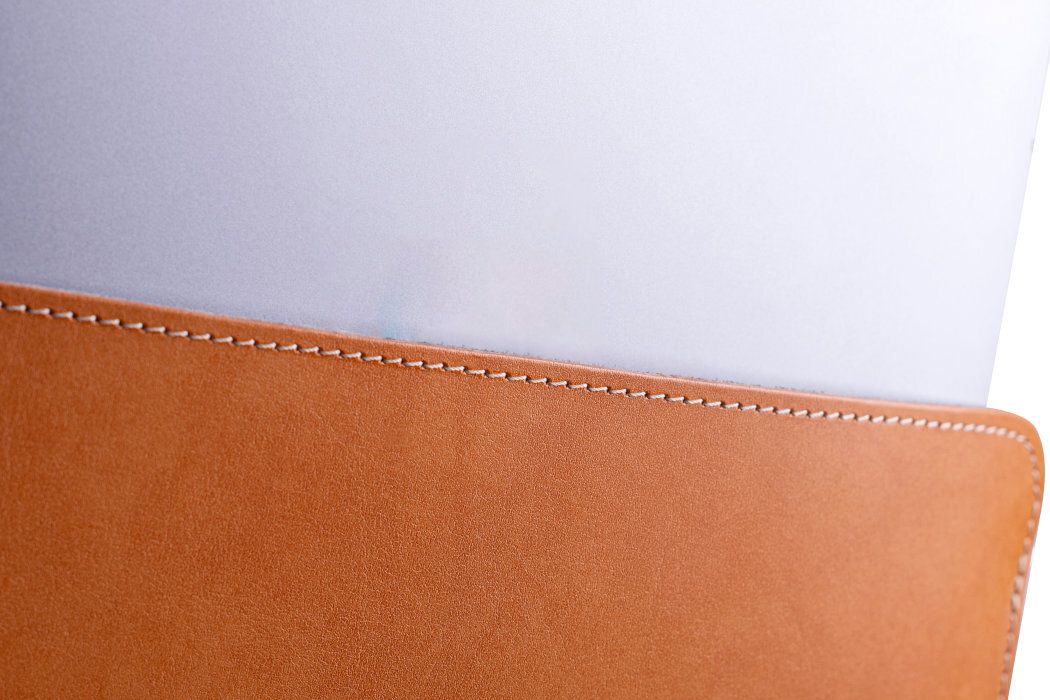 Obraz przedstawia brązowe etui na laptopa z widocznym białym szwem na krawędzi. W etui znajduje się srebrny laptop, którego część wystaje ponad krawędź etui.