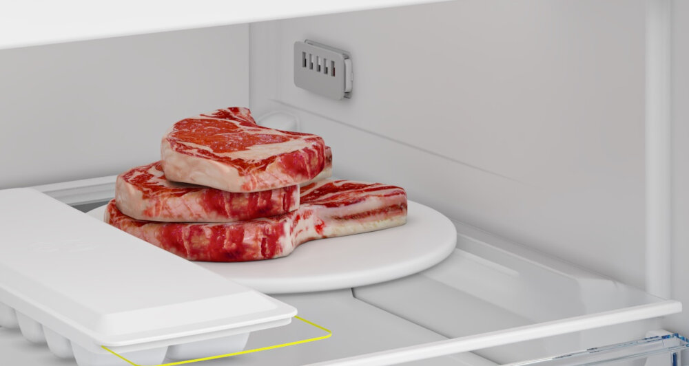 ZAMRAŻARKA BEKO B5RFNE274W Zbliżenie na górną półkę zamrażarki z tacą do kostek lodu oraz świeżym mięsem przeznaczonym do zamrożenia. Grafika prezentuje przestrzeń do szybkiego schładzania i przechowywania produktów spożywczych alarm drzwi 232 kWh klasa E oszczędność energii bezpieczeństwo żywności