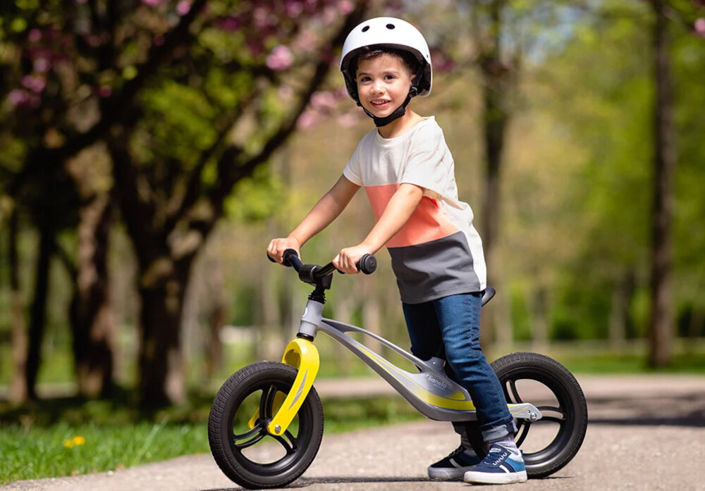 Rowerek biegowy LIONELO Bart Tour Szaro-żółty park dziecko siedzi na rowerku w tle drzewa i rośliny dróżka rama w technologii MONOCOQUE stworzona z pojedynczego odlewu stopu magnezu o 75% lżejszy od stali wysoka odpornością na uszkodzenia połączenie niskiej wagi 3,3 kg z wytrzymałością komfort i bezpieczeństwo