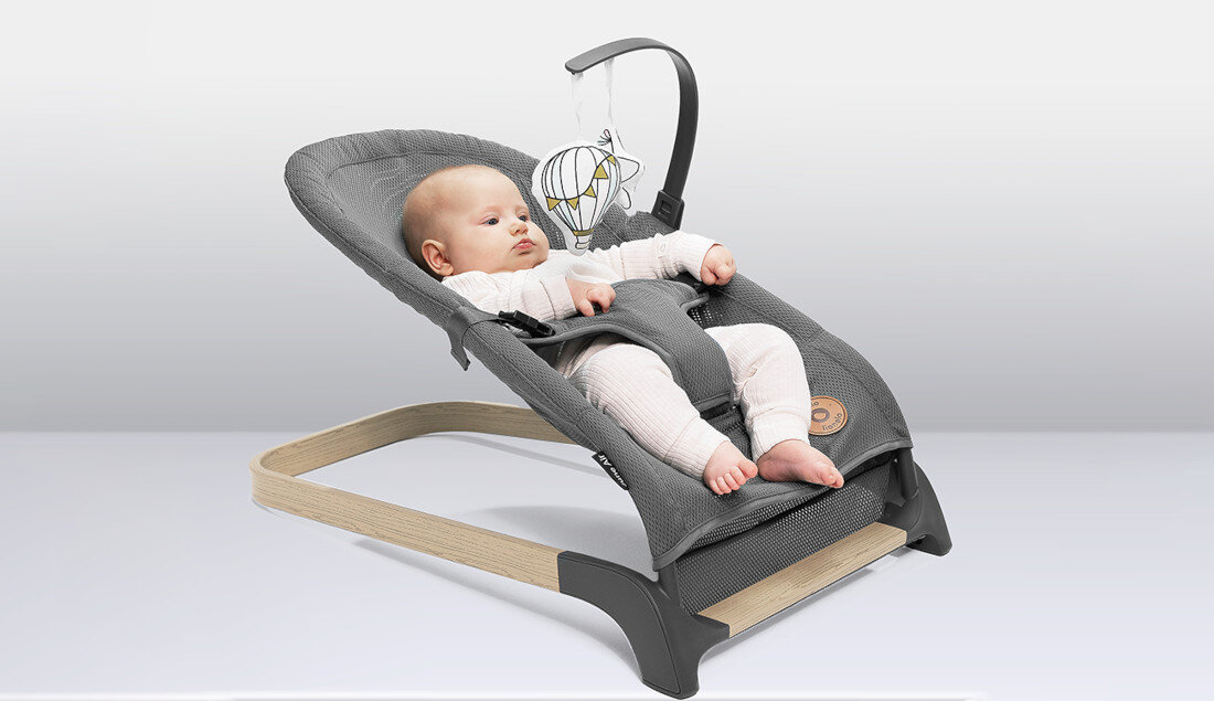 Na obrazie widoczny jest szary bujaczek z drewnianą podstawą, w którym leży niemowlę. Nad bujaczkiem zawieszona jest zabawka w kształcie piłki.