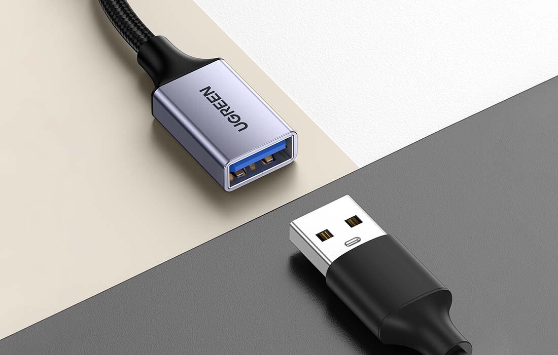 Na obrazie widoczny jest adapter USB-C/USB-A z napisem \'UGREEN\' na obudowie oraz kabel USB z wtyczką. Tło jest w odcieniach beżu i szarości.