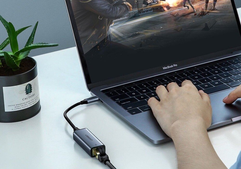 Adapter USB-C - RJ-45 UGREEN CM199 Dłoń osoby na klawiaturze srebrnego laptopa, do którego boku podłączony jest szary adapter USB-C z wpiętym kablem sieciowym Ethernet, a obok stoi mała czarna doniczka z aloesem