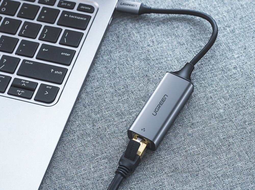 Adapter USB-C - RJ-45 UGREEN CM199 Zbliżenie na szary adapter z napisem 'UGREEN' podłączony krótkim przewodem USB-C do boku srebrnego laptopa, z wpiętym od dołu czarnym kablem sieciowym na tle szarej tkaniny