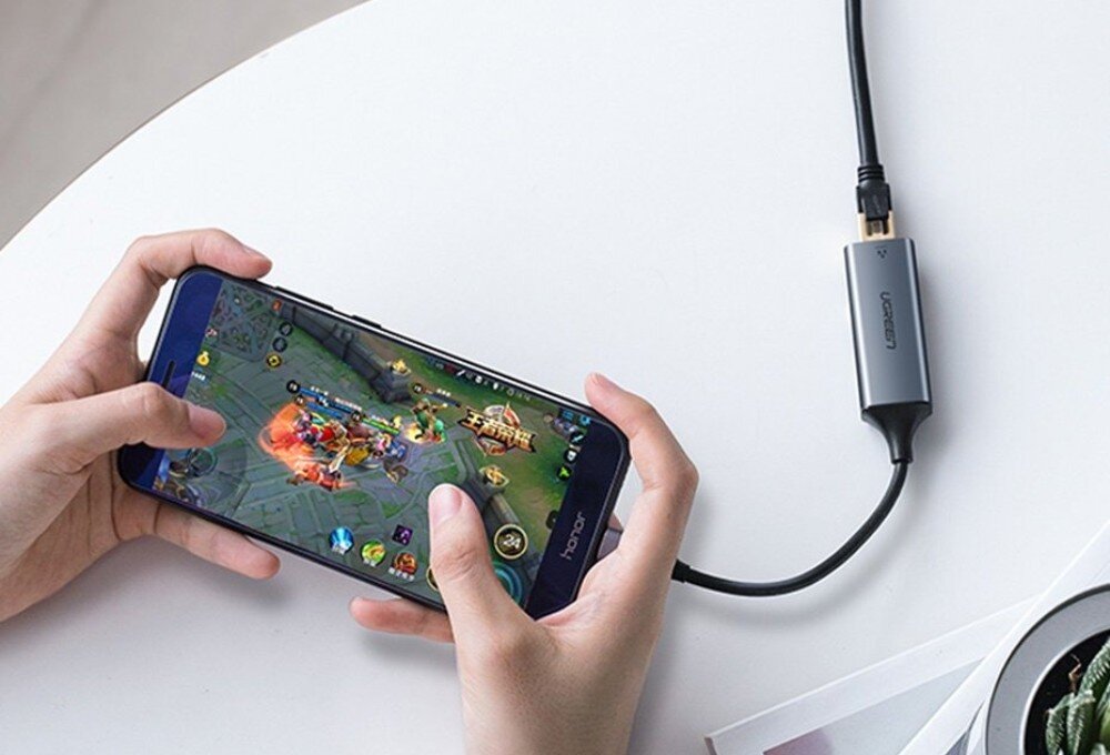Adapter USB-C - RJ-45 UGREEN CM199 Osoba trzymająca w dłoniach smartfon z włączoną grą mobilną, do którego przez port USB-C podłączony jest szary adapter z wpiętym kablem sieciowym, leżący na białym blacie