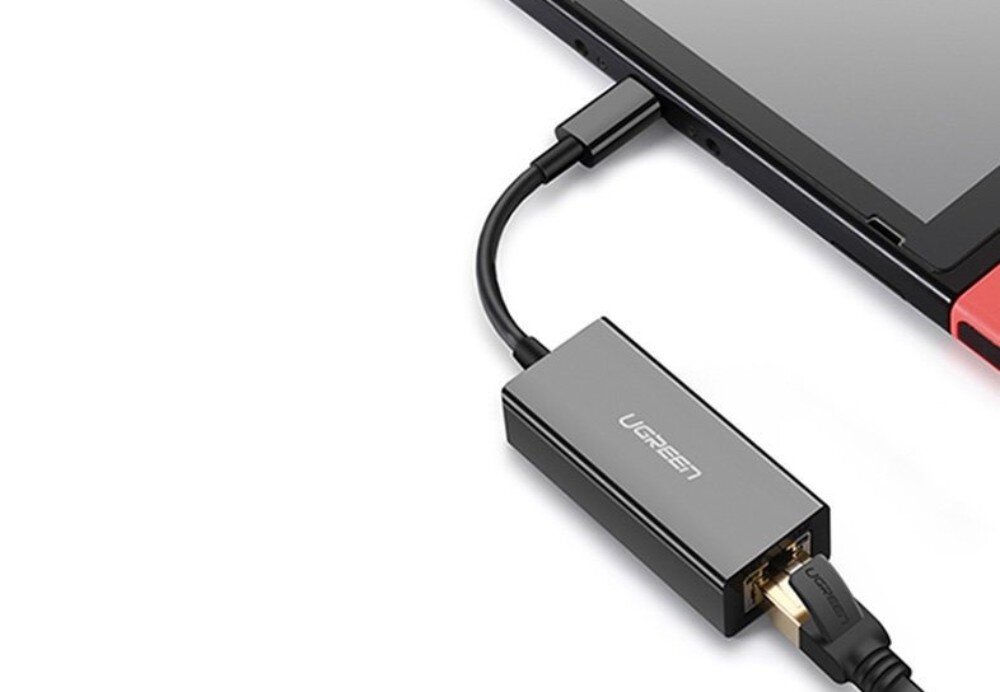 Adapter USB-C - RJ-45 UGREEN CM199 Szary adapter podłączony przewodem USB-C do dolnej części konsoli przenośnej z widocznym czerwonym kontrolerem, z wpiętym od spodu kablem sieciowym Ethernet na białym tle