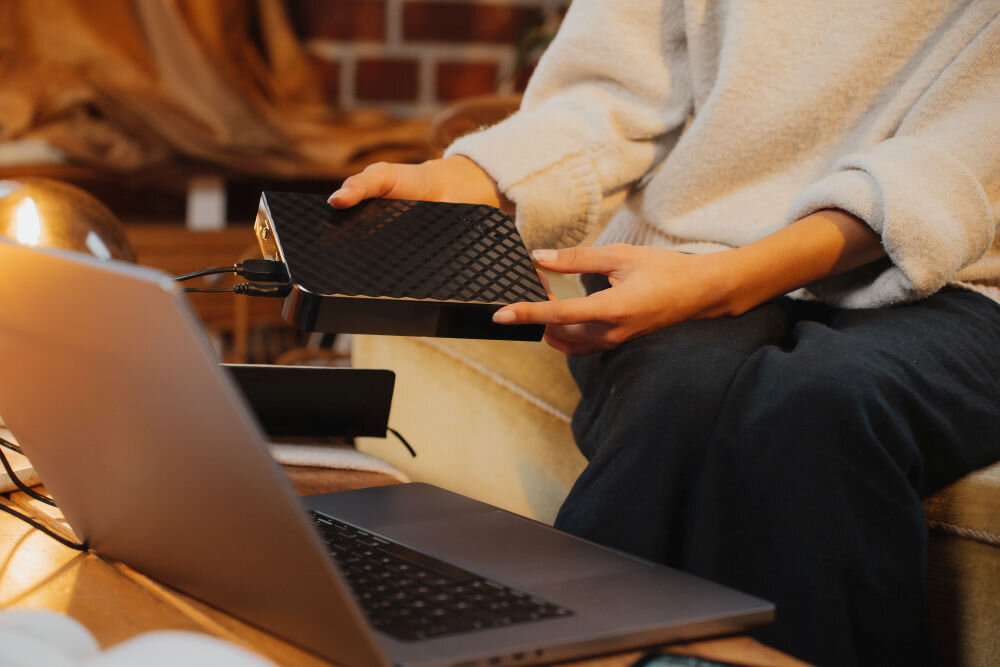 Osoba trzyma czarny dysk zewnętrzny o fakturze kratki, podłączony do laptopa na stole. W tle widoczne są elementy wnętrza, takie jak kanapa i ceglana ściana.