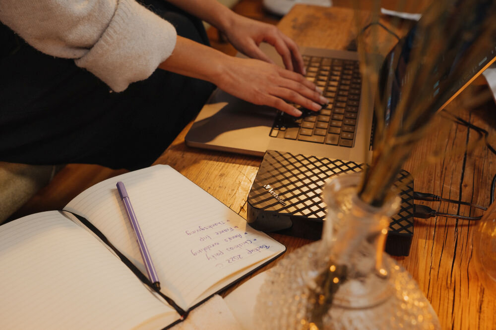 Osoba pisze na laptopie, obok którego znajduje się czarny dysk zewnętrzny z logo \'VERBATIM\'. Na stole leży otwarty notes z długopisem i odręcznymi notatkami.