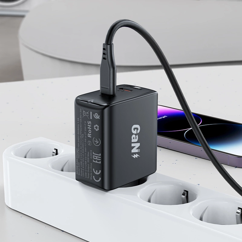 Czarna szybka ładowarka sieciowa GaN 35W z dwoma portami USB-C jest podłączona do białej listwy zasilającej. Obok leży smartfon z widocznym ekranem.