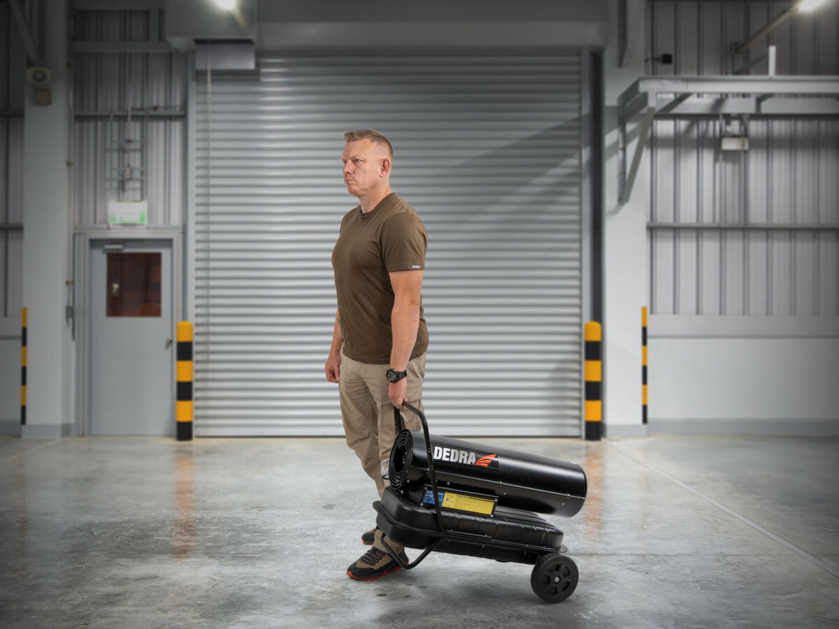 Nagrzewnica olejowa DEDRA DED9952B, koła, uchwyt, ramka, wytrzymałość, Mężczyzna ciągnie nagrzewnicę Dedra DED9952B na kołach w przestronnej hali przemysłowej.