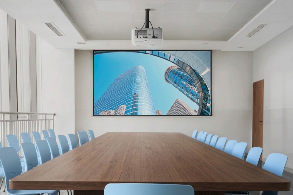 Projektor laserowy VIEWSONIC Nowoczesna sala konferencyjna z długim drewnianym stołem i krzesłami, z ekranem wyświetlającym wieżowce oraz podwieszonym pod sufitem projektorem