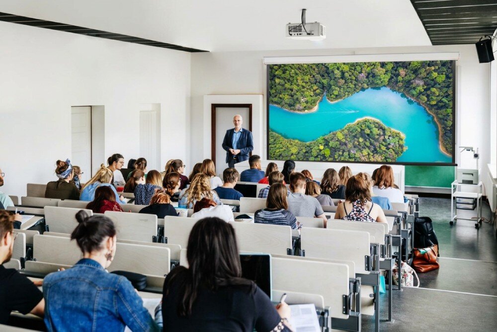 Projektor laserowy VIEWSONIC Sala wykładowa ze studentami siedzącymi w ławkach, wykładowcą przy oknie oraz ekranem wyświetlającym widok z góry na rzekę i las
