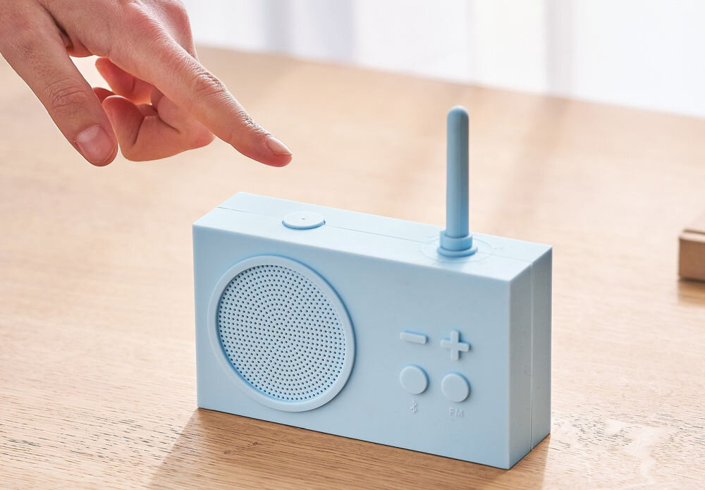 Jasnoniebieski radioodtwarzacz z widocznym głośnikiem, anteną i przyciskami do regulacji głośności oraz symbolami Bluetooth i FM. Dłoń zbliża się do przycisku na górze urządzenia.
