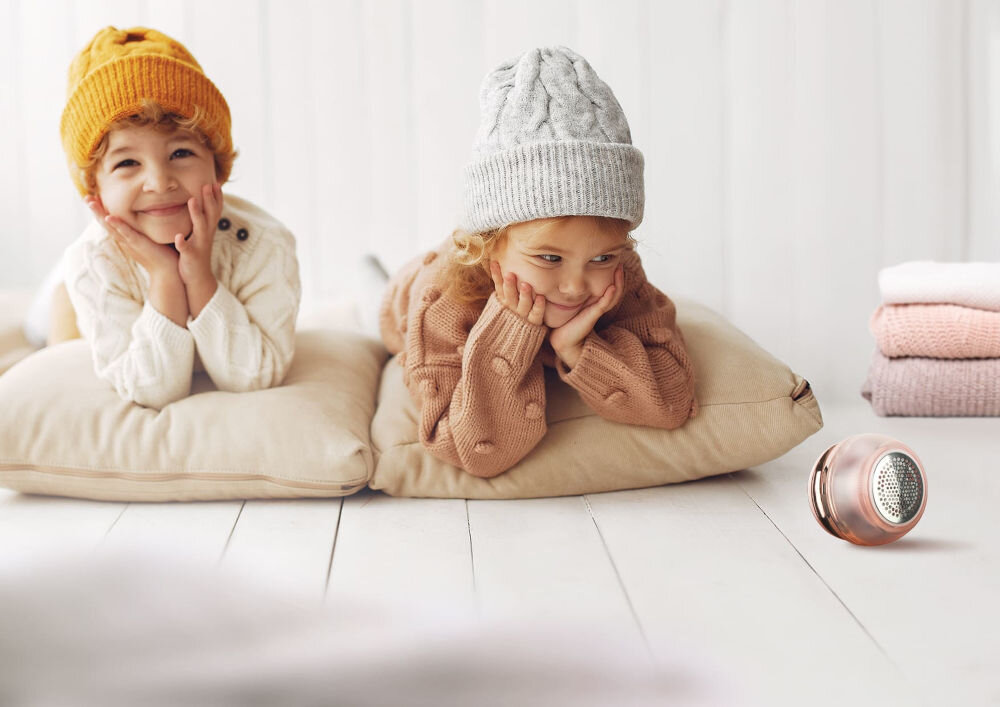 Golarka do ubrań NENO Merino Dwoje uśmiechniętych dzieci w zimowych czapkach i swetrach leży na podłodze obok golarki do ubrań Neno.