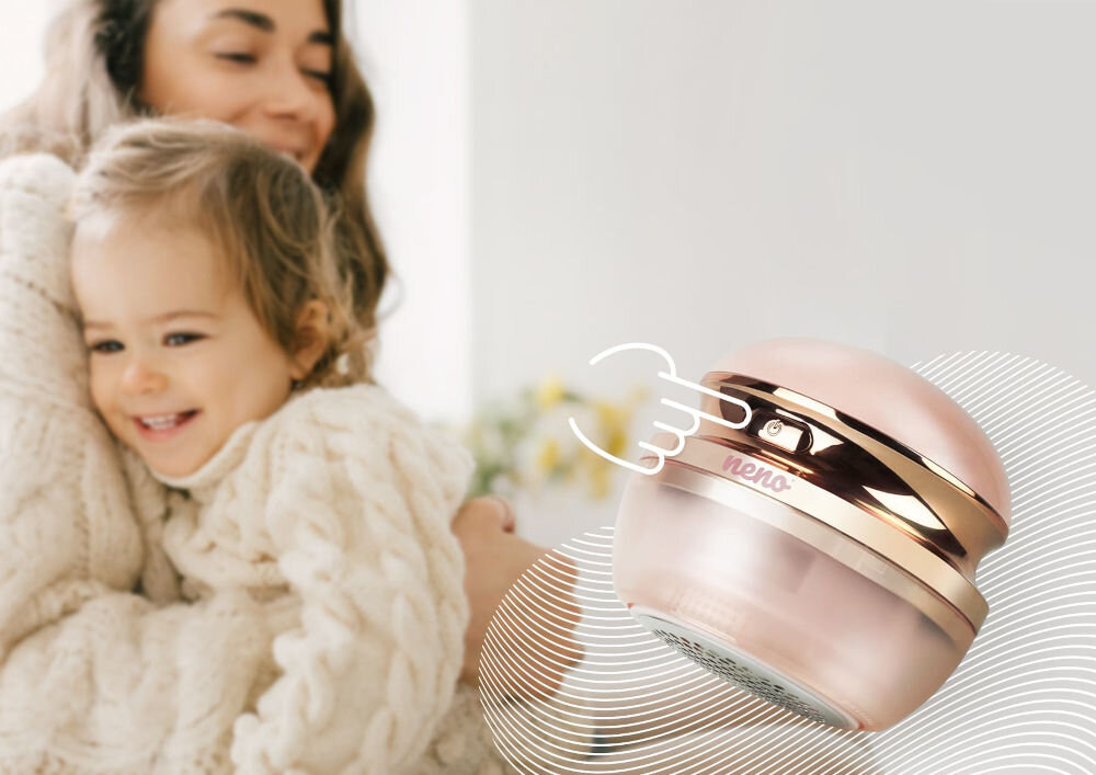 Golarka do ubrań NENO Merino Zbliżenie na różową golarkę do ubrań Neno z widocznym przyciskiem zasilania, w tle rozmazana postać uśmiechniętej kobiety z dzieckiem.